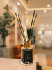 Luxury reed diffuser by Nana’s Fragrance with black reeds in a clear glass bottle, placed on a white surface with a softly lit, elegant interior background.